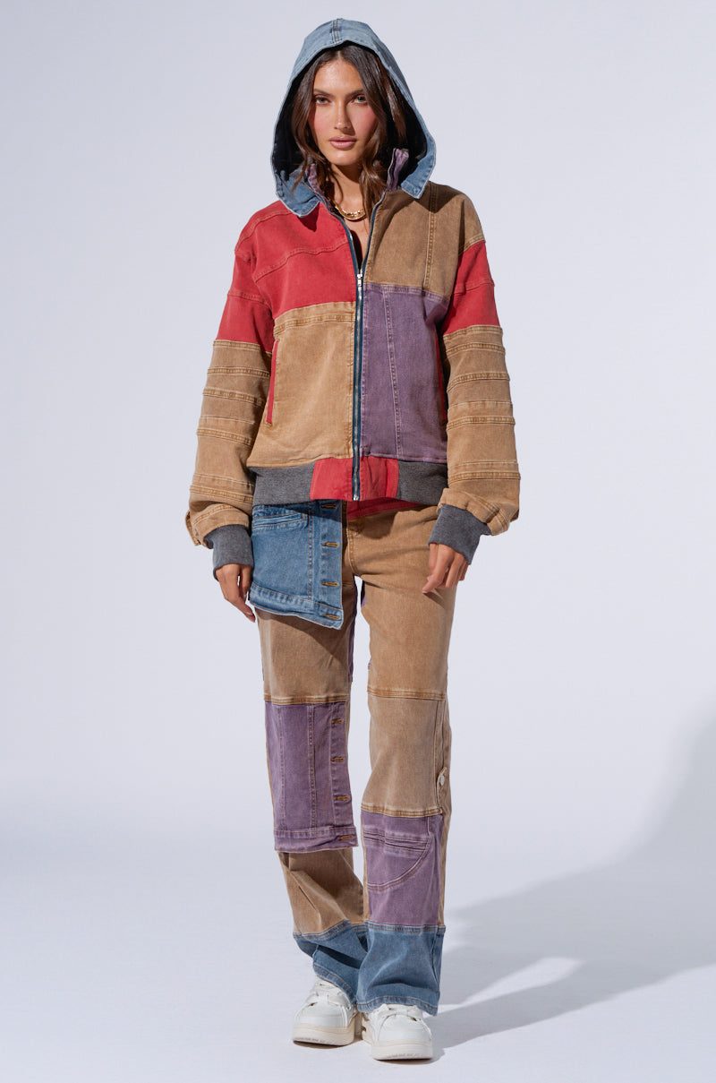 A person stands against a plain background, wearing a hooded, patchwork jacket in shades of brown, red, and purple by AKIRA Label. They are paired with matching SUPERNATURAL PATCHWORK JEANS featuring a high rise waist. They also sport white sneakers and have their hands in the jacket pockets, with the hood pulled up.
