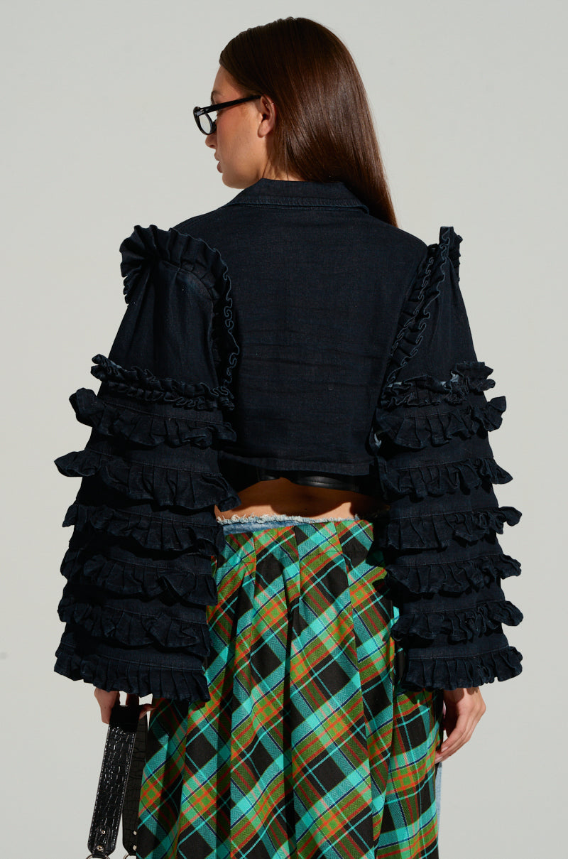 A rear view of a person dressed in the FELICITY STATEMENT SLEEVE DENIM JACKET features statement sleeves and ruffle trim. They have long brown hair, wear glasses, and carry a small handbag. The skirt displays a green and orange plaid pattern against the plain gray background.