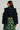 A rear view of a person dressed in the FELICITY STATEMENT SLEEVE DENIM JACKET features statement sleeves and ruffle trim. They have long brown hair, wear glasses, and carry a small handbag. The skirt displays a green and orange plaid pattern against the plain gray background.