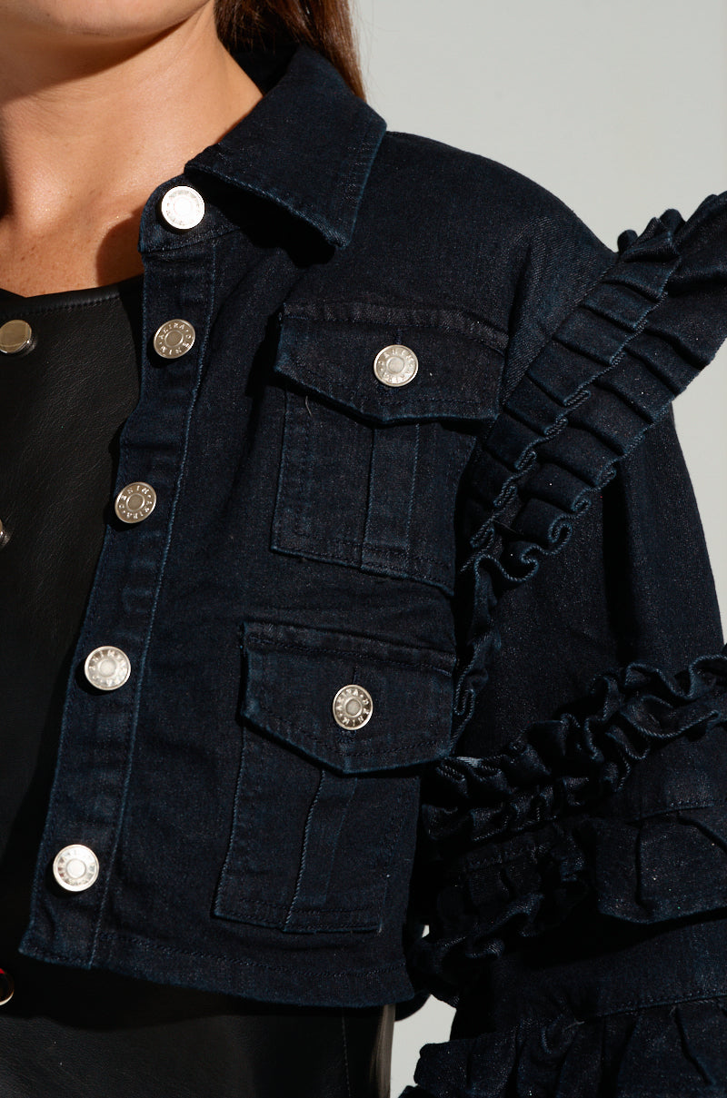A close-up shows someone wearing the FELICITY STATEMENT SLEEVE DENIM JACKET, characterized by its dark denim fabric, silver buttons, and ruffle trim on the eye-catching sleeves. The jacket is styled with flap pockets and is layered over a black top.