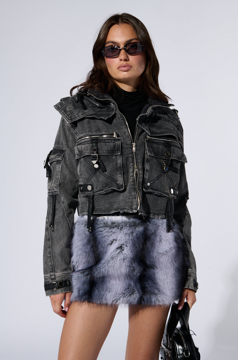 A person with long brown hair wears dark sunglasses, a black turtleneck, a SCORPIO CARGO DENIM JACKET featuring multiple pockets and metallic zipper detailing, and a fluffy gray skirt. They are holding a black handbag with faux leather strap detailing and standing against a plain light background. 3