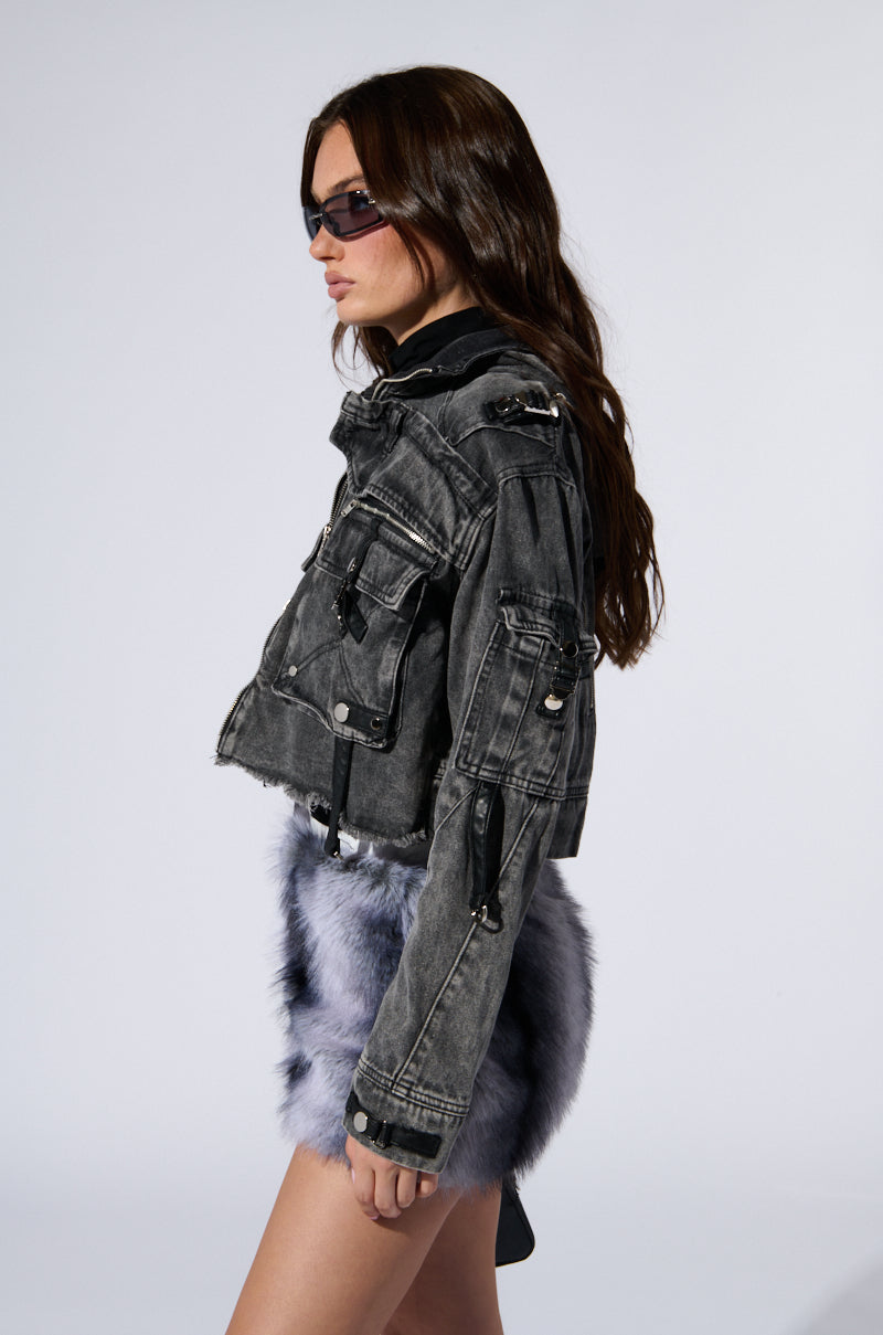 A person with long dark hair, wearing sunglasses, a SCORPIO CARGO DENIM JACKET characterized by its multiple zippers and pockets along with faux leather strap detailing, and a blue furry skirt stands against a plain light background. They are facing slightly to the left. 4