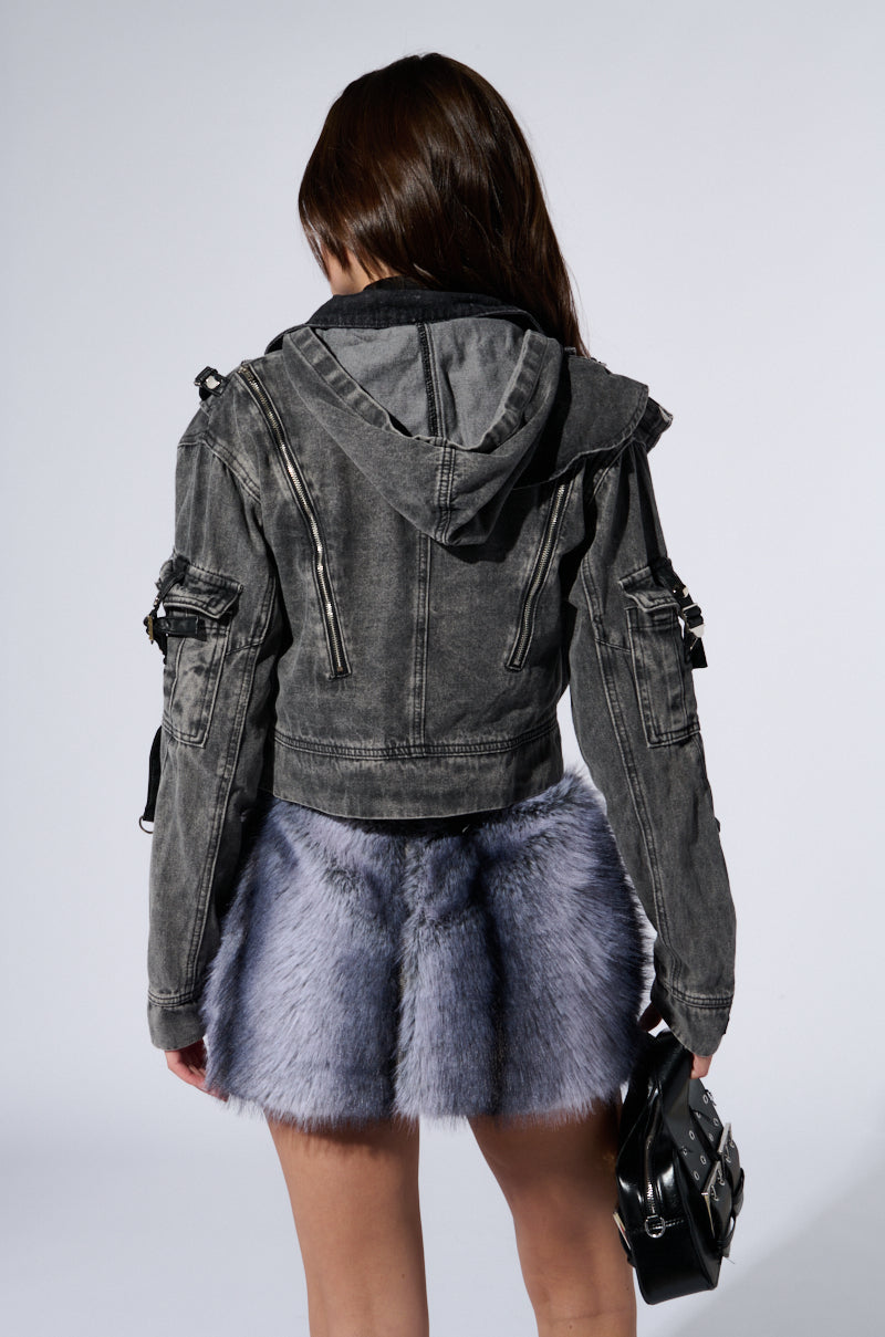 A person stands with their back to the camera, wearing a SCORPIO CARGO DENIM JACKET featuring metallic zipper detailing and a short, fluffy purple skirt. They hold a small black bag with faux leather strap detailing in their right hand, and their long hair cascades down their back against a plain, light-colored background. 5