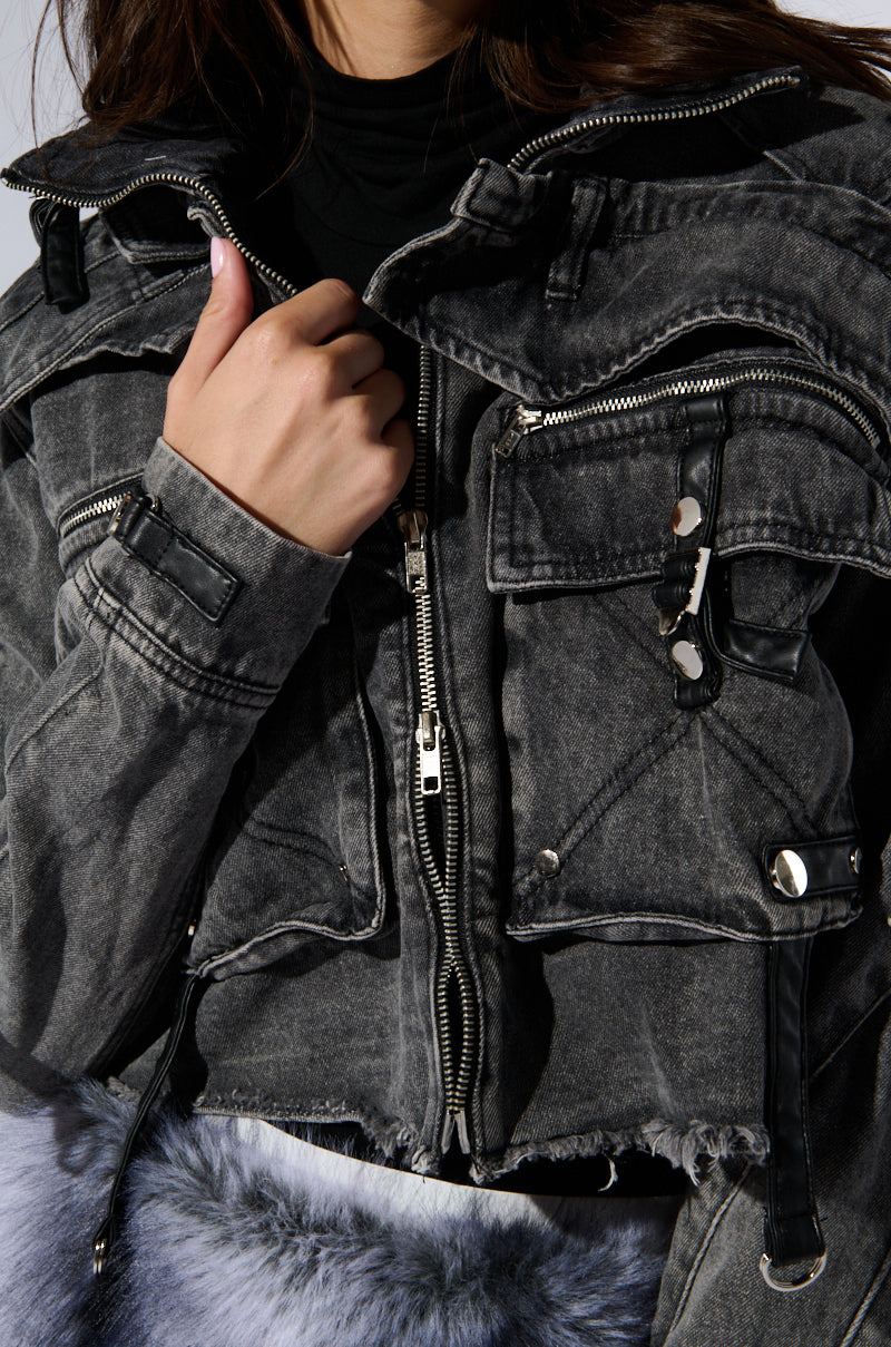 A person wearing a black, distressed SCORPIO CARGO DENIM JACKET with multiple zippers and pockets is pictured. The jacket, featuring metallic zipper detailing, is partially unzipped to reveal a black shirt underneath. The lower part of the face and upper body are visible as hands adjust the jacket. 8