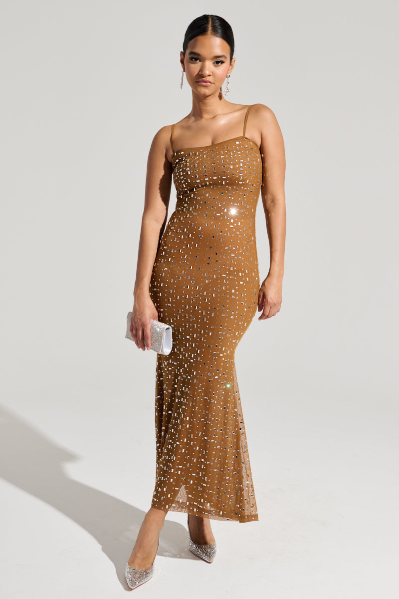 The individual is dressed in the ANASTASIA MESH RHINESTONE MAXI DRESS, featuring a curve-hugging fit and adjustable cami straps adorned with sequins. They complement the look with a small silver clutch and silver high heels, standing out against the plain gray background that highlights their elegant attire.