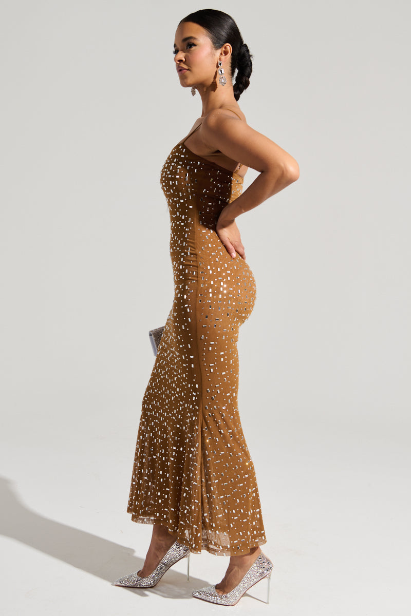 A woman stands in profile wearing the ANASTASIA MESH RHINESTONE MAXI DRESS, featuring a curve-hugging fit and decorated with white polka dots. She complements the dress with sparkling high-heeled shoes and dangling earrings. Her hair is styled in a neat bun, and she holds a small purse by her side.