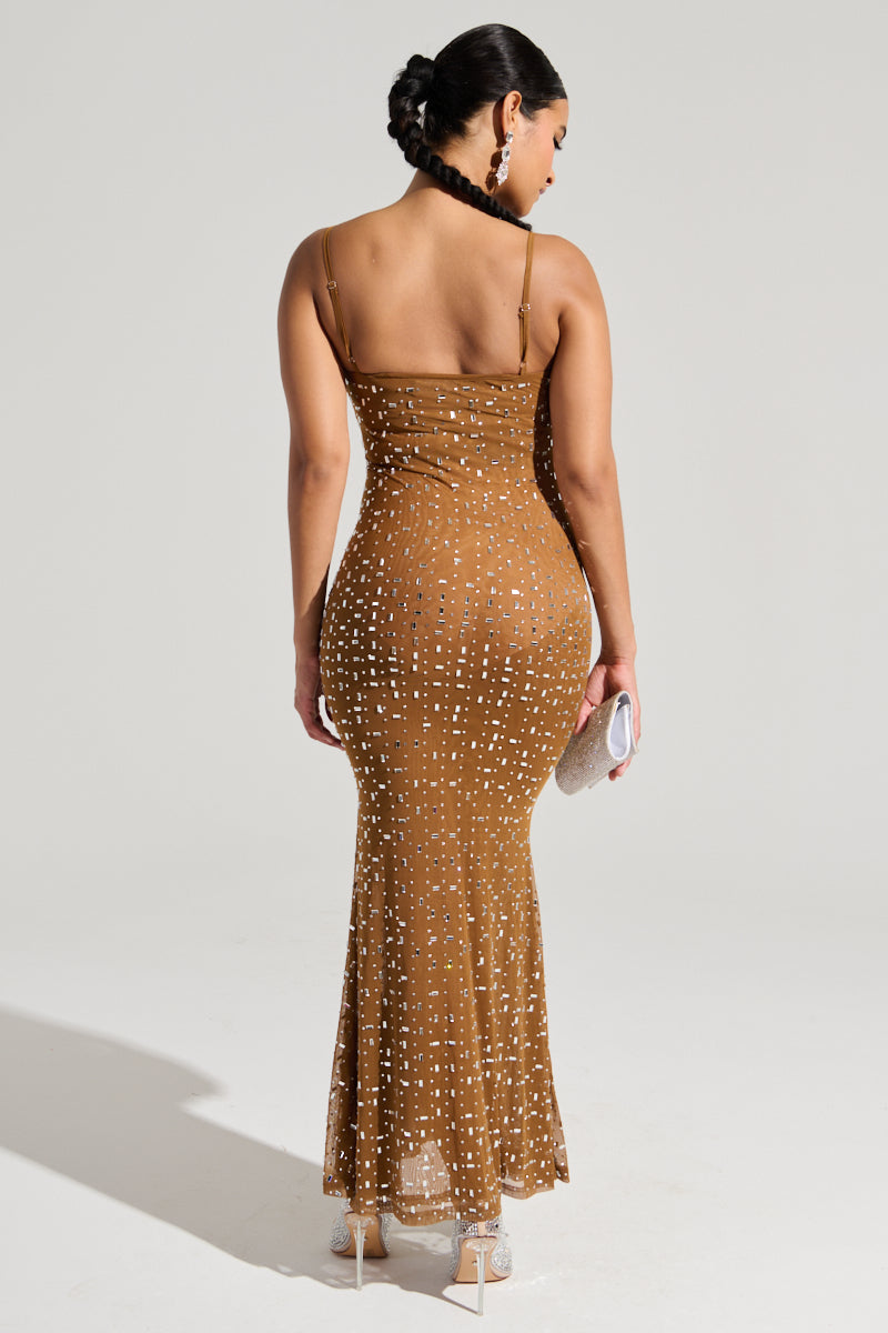 A woman in the ANASTASIA MESH RHINESTONE MAXI DRESS, featuring a curve-hugging fit and white polka dots on a brown fabric, holds a small clutch. Her hair is styled in an elegant updo, and she wears high heels with adjustable cami straps, standing against a neutral background.