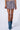 A person stands against a plain background wearing the ALEXANDRA PLEATED MINI SKIRT with a textured pattern and distressed hem, paired with mid-calf denim-inspired knit boots. The image focuses on the lower half of the body, highlighting the high-rise design of the skirt.