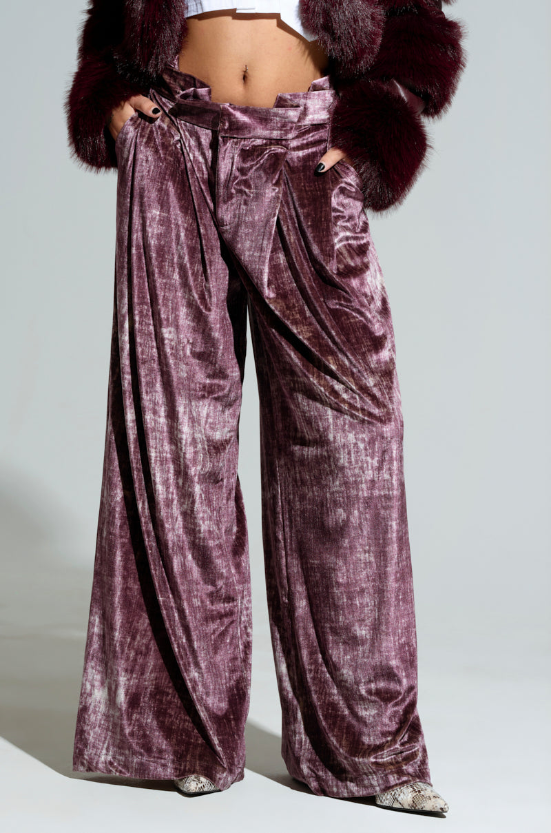 A person wearing the TALK ABOUT ME TROUSER, featuring velvet-inspired, wide-legged purple pants and a cropped jacket with fur sleeves, posing with hands in pockets. The photo highlights the lower body, showcasing the luxurious fabric and loose fit of the wide-leg silhouette.