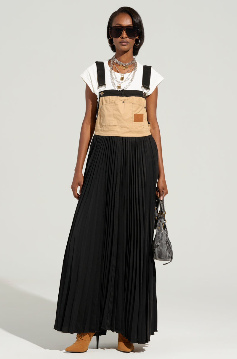 A woman showcases an effortlessly chic look against a plain light gray background, featuring the FOR THE STREETS OVERALL MAXI DRESS worn over a white t-shirt. Her ensemble includes oversized sunglasses, a long black pleated skirt, and layered necklaces. She accessorizes with a small black bag and tan boots.