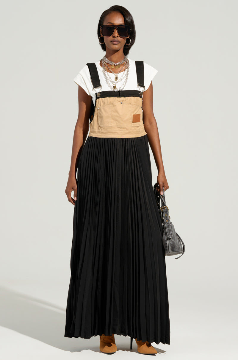 A person sporting sunglasses, a white t-shirt, and the FOR THE STREETS OVERALL MAXI DRESS, which highlights a distinctive beige apron-like top with black straps paired with a long pleated black skirt. They carry a small black bag and layer multiple necklaces while standing against a plain background.