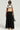 A person sporting sunglasses, a white t-shirt, and the FOR THE STREETS OVERALL MAXI DRESS, which highlights a distinctive beige apron-like top with black straps paired with a long pleated black skirt. They carry a small black bag and layer multiple necklaces while standing against a plain background.