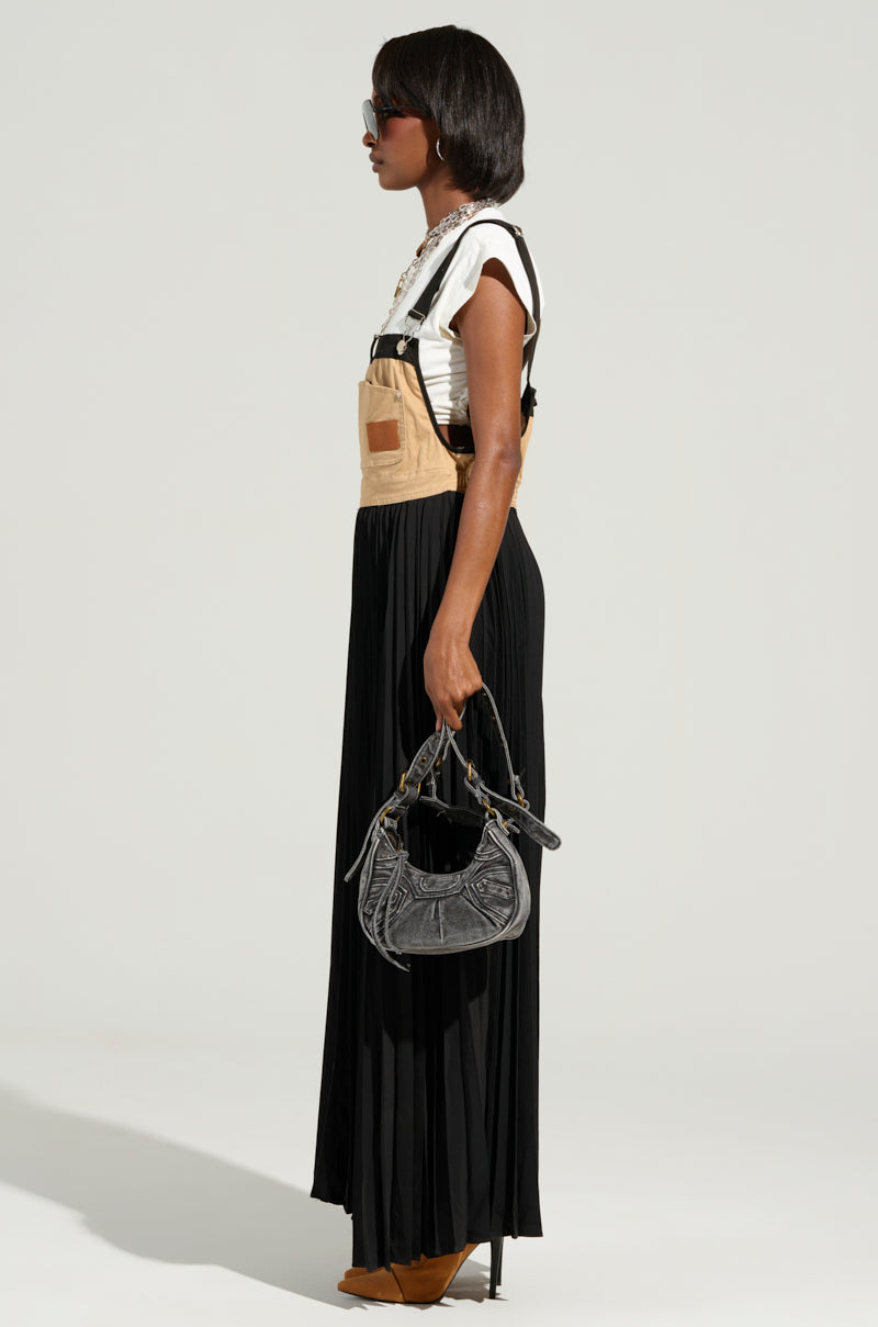 A woman stands in profile, wearing stylish sunglasses and the FOR THE STREETS OVERALL MAXI DRESS—featuring a white cotton knit base, beige overalls, and a long black pleated skirt. She complements her outfit with a black handbag and tan high-heeled shoes. The neutral background enhances her utility-inspired style.
