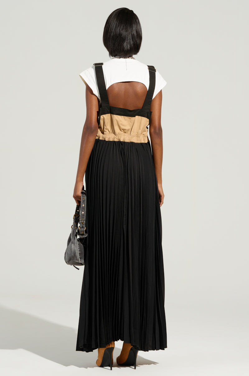 A person with short black hair is wearing a white top and the FOR THE STREETS OVERALL MAXI DRESS, a sleeveless, utility-inspired black and beige dress with a pleated skirt. They hold a black handbag while standing against a neutral background.