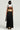 A person with short black hair is wearing a white top and the FOR THE STREETS OVERALL MAXI DRESS, a sleeveless, utility-inspired black and beige dress with a pleated skirt. They hold a black handbag while standing against a neutral background.
