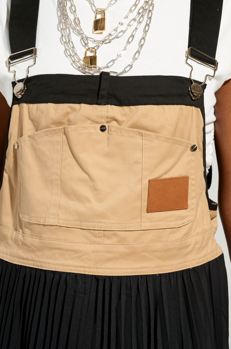 The individual sports a utility-inspired style, wearing a white shirt and layered silver chains beneath the FOR THE STREETS OVERALL MAXI DRESS, which features large pockets and a brown patch, perfectly paired with a black pleated skirt.