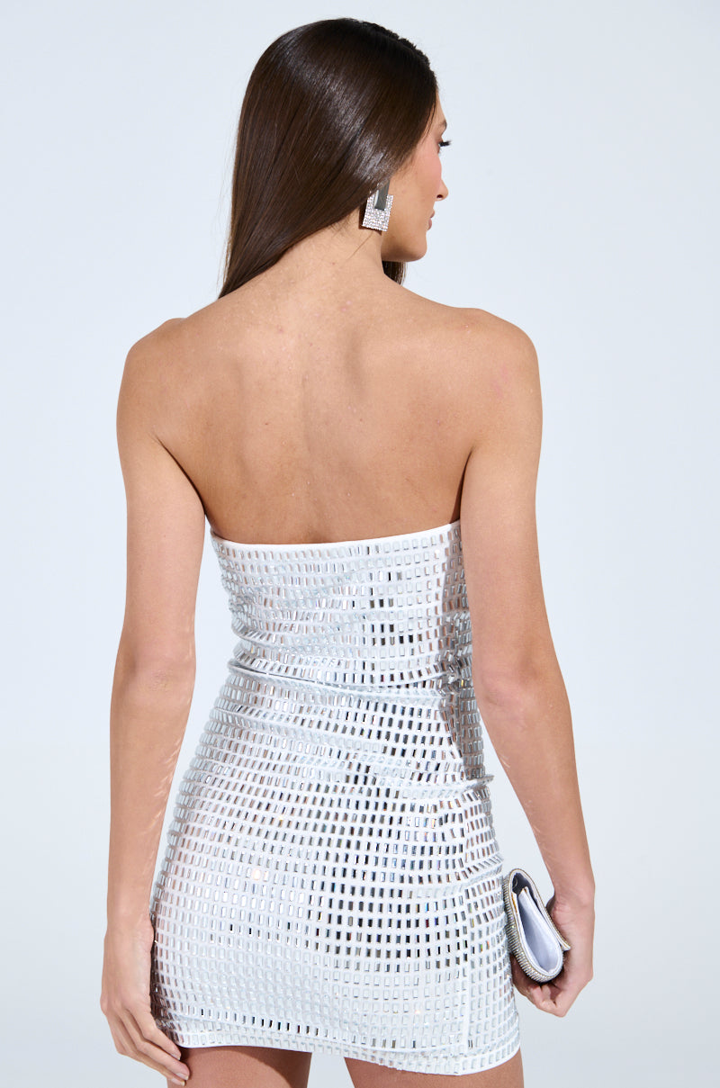 A woman with long brown hair is wearing a sparkling, strapless "MEET ME ON THE DANCE FLOOR" rhinestone mini dress in white and large square earrings. She holds a silver clutch with her back to the camera against a plain light gray background.