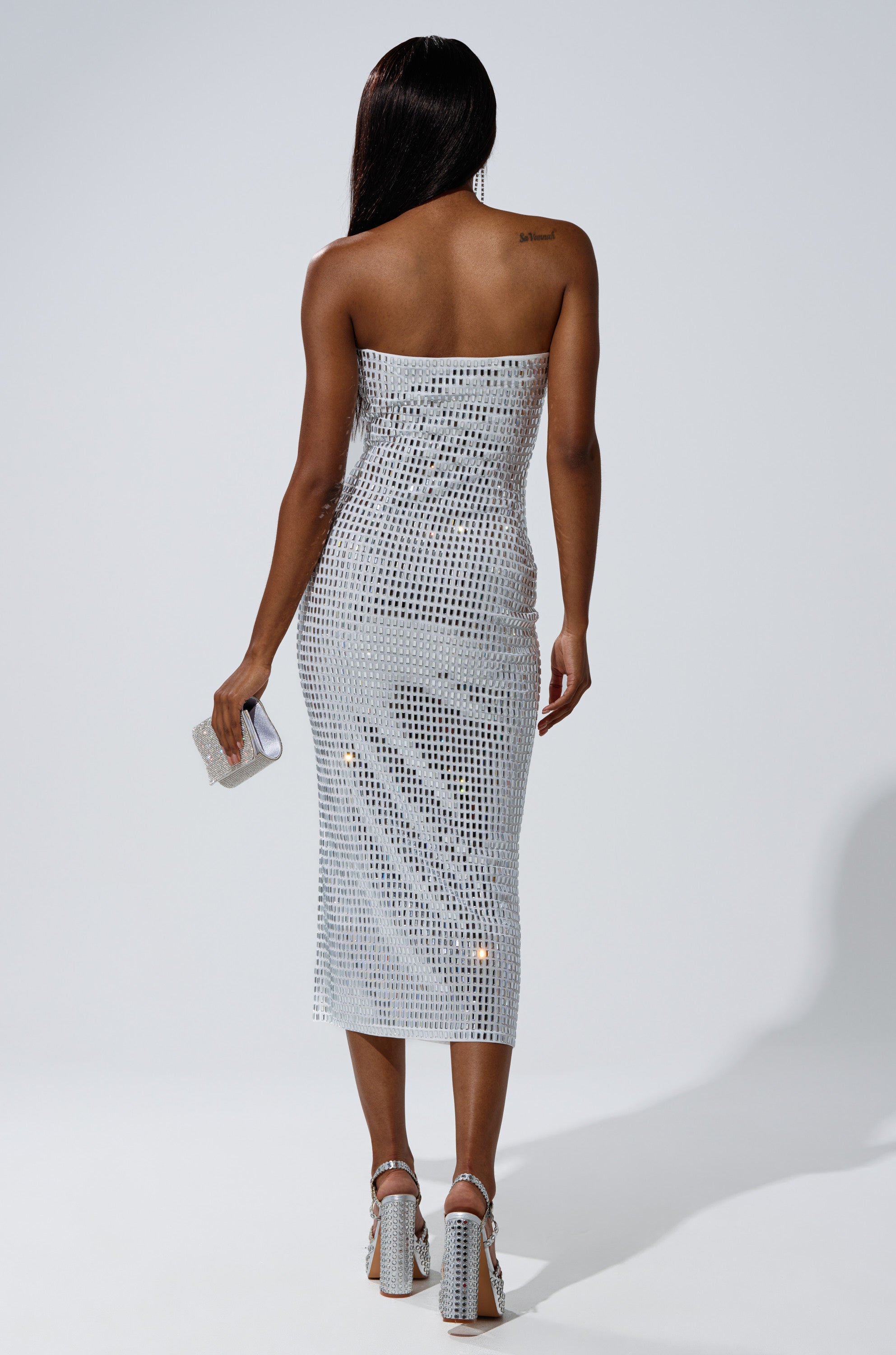 A woman faces away, wearing the CAN'T SHINE BRIGHTER THAN ME RHINESTONE MIDI DRESS IN WHITE with silver sequins. She holds a small silver clutch, has long dark hair, and wears high-heeled sandals with clear straps.
