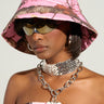 A person wearing a YOU CANT SEE ME CAMO BUCKET HAT and strapless top, complemented by sleek sunglasses and large hoop earrings. A chunky metallic choker and chain necklace with a heart-shaped pendant complete the bold fashion look.