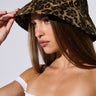 A woman wearing the WILD ONE CHEETAH BUCKET HAT adjusts its brim. She has long straight hair, is wearing an off-the-shoulder white cotton top, and is looking to the side with a neutral expression. The background is plain and light-colored.