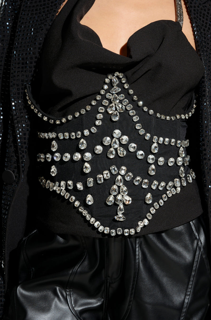 A person is dressed in a black outfit featuring a sheer mesh top with structured draping and shimmering embellishments, paired with black leather pants. The ensemble is adorned with intricate patterns of shiny, faceted gems and is complemented by the JODIE RHINESTONE BELT that adds an extra touch of sparkle.