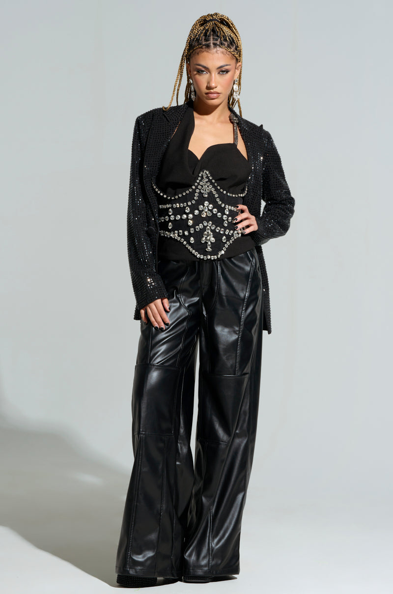 A person with braided hair is wearing a sequined black jacket and a detailed corset-style top, paired with wide-leg leather pants. They stand confidently with one hand on their hip, showcasing the JODIE RHINESTONE BELT's underbust design against a plain background.