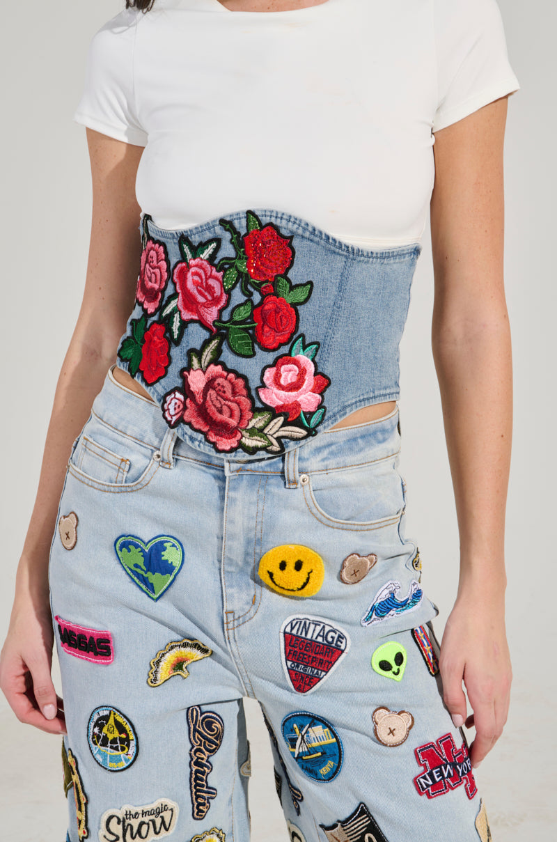 A person wears a white t-shirt with blue jeans featuring the ROSE NIGHT PATCH BELT, highlighted by embroidered pink and red roses. The jeans are decorated with colorful patches including hearts, skulls, and various logos.
