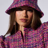 A person with long hair is wearing a fashionable TWEED PRINCESS BUCKET HAT and a matching jacket, both adorned in vibrant shades of pink, purple, and yellow. The outfit showcases a woven, tweed-like multicolor pattern with metallic details that add a hint of sparkle. They look directly at the camera against a plain background.