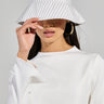 A person embodies a casual look with a white, long-sleeved top and the DELILA PINSTRIPE BUCKET HAT, which partially covers their face. Large hoop earrings enhance their summer style against a neutral background.