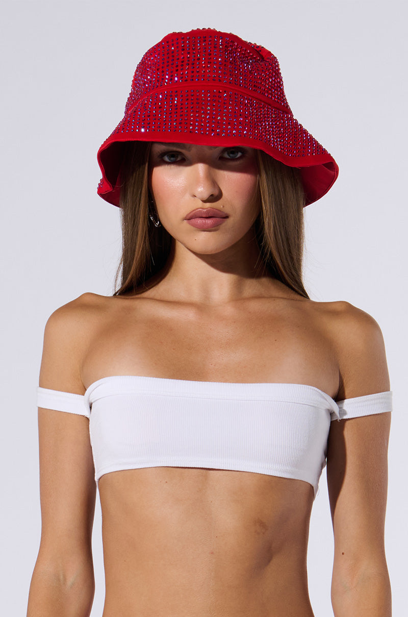 A woman with long hair is wearing an ON GO BLING BUCKET HAT in red, adorned with rhinestone embellishments and a white, off-shoulder, bandeau-style crop top. She has a neutral expression and stands against a plain grey background.