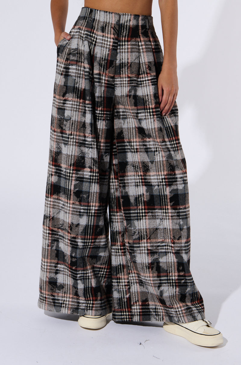 A person is styled in the PLAID SWAGGER SLACKS, which boast a high-rise waist and a wide-leg silhouette in a classic blend of black, gray, and brown tones. Their hands are casually tucked into the pockets of these chic trousers. They complete the look with white sneakers against a plain white backdrop.