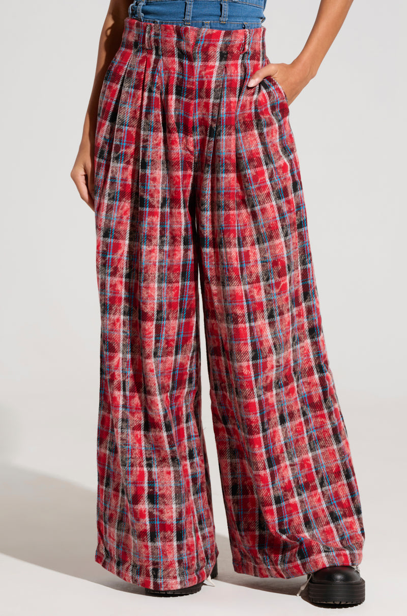 A person wearing PLAID SWAGGER SLACKS, a pair of high-rise, wide-leg pants with red, black, and white plaid patterns. The slacks feature pleats and the individual has one hand in their pocket. They are also sporting a blue top and black shoes against a plain white background.