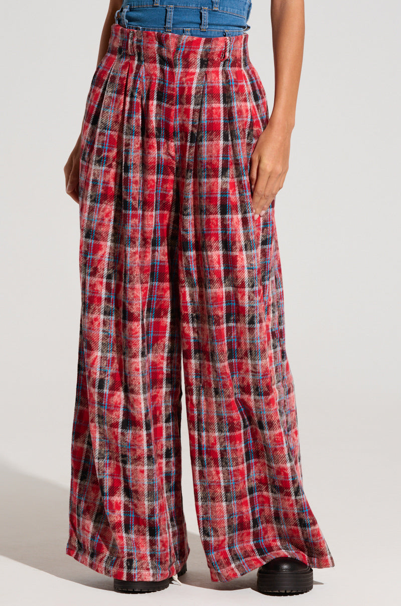 A person wearing the PLAID SWAGGER SLACKS, which are high-waisted, wide-leg pants in a red, black, and white plaid pattern. These cotton slacks have pleats at the waist and are cinched with a belt. They are paired with black shoes. Only the lower torso and legs are visible.