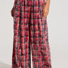 A person wearing the PLAID SWAGGER SLACKS, which are high-waisted, wide-leg pants in a red, black, and white plaid pattern. These cotton slacks have pleats at the waist and are cinched with a belt. They are paired with black shoes. Only the lower torso and legs are visible.