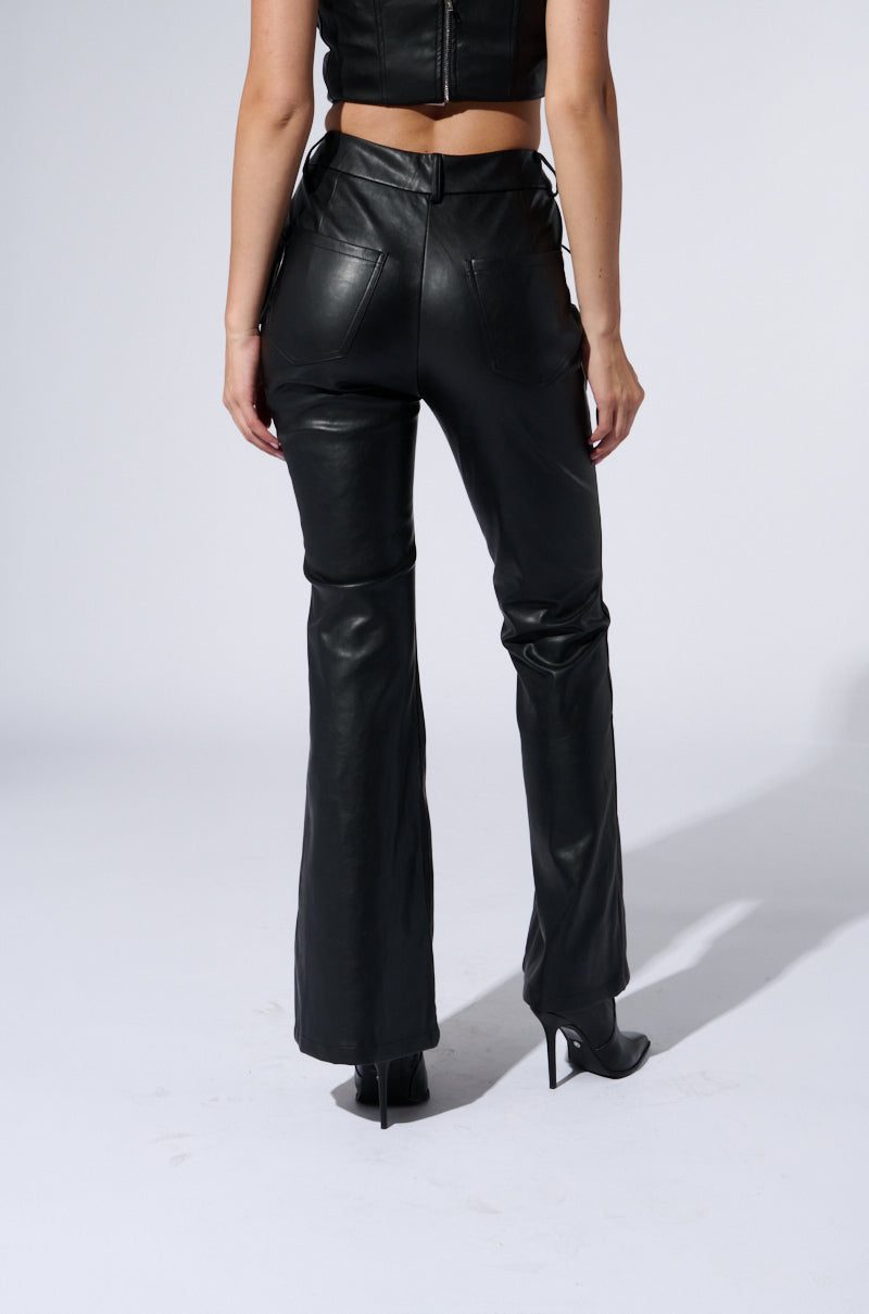 A person stands facing away, shown from the lower back down. They are wearing H-TOWN HOTTIE FAUX LEATHER LACE UP PANT and black high-heeled boots, paired with a black top from the AKIRA Label. The background is plain and light-colored.