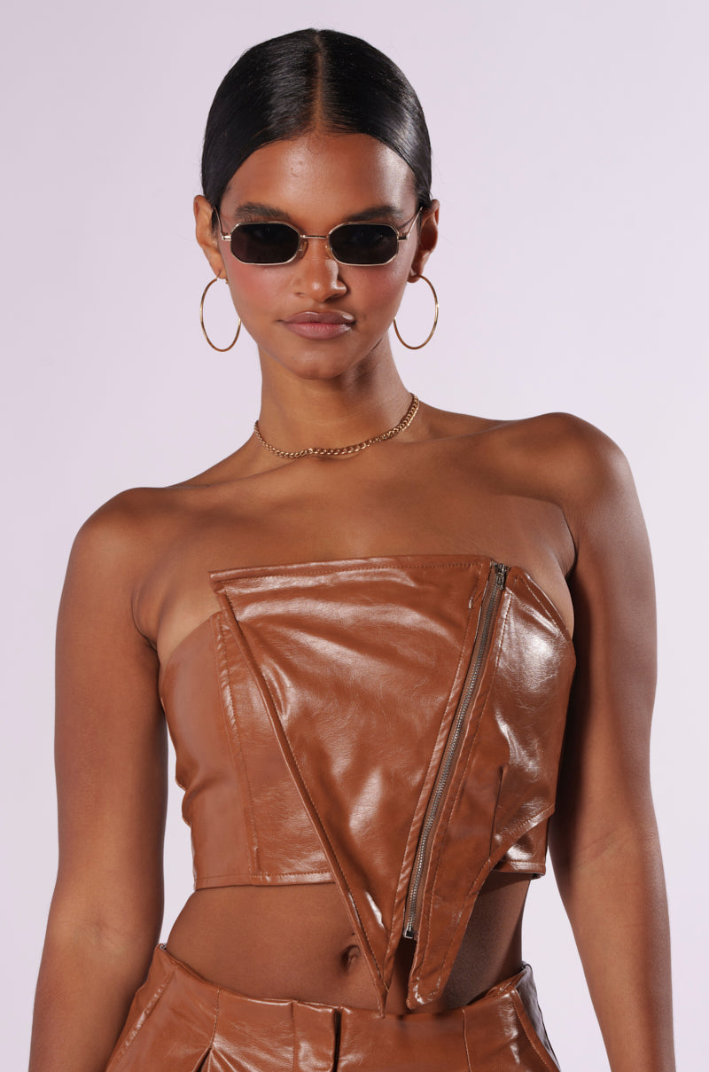 A person with dark hair styled in a neat updo wears small, oval-shaped sunglasses, large hoop earrings, and the LOOK BACK AT IT CORSET in brown faux leather with an off-shoulder design and an asymmetrical zipper. They also have a gold chain necklace against a plain white background. 0