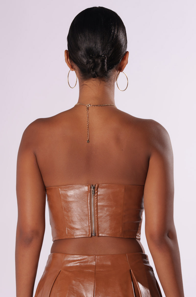 A person is seen from the back, wearing a LOOK BACK AT IT CORSET in a brown, strapless faux leather design with a zipper closure, paired with matching brown leather pants. Their hair is styled in a low bun, and they are accessorized with hoop earrings and a delicate necklace featuring a small dangling charm. 3
