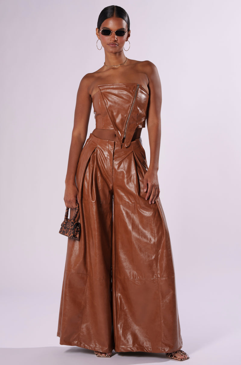 Woman wearing a brown faux leather strapless top and wide-leg trousers, holding a small brown patterned handbag. She has accessorized with dark sunglasses, large hoop earrings and looks effortlessly chic in the LOOK BACK AT IT CORSET from the AKIRA Label, standing against a plain background. 4