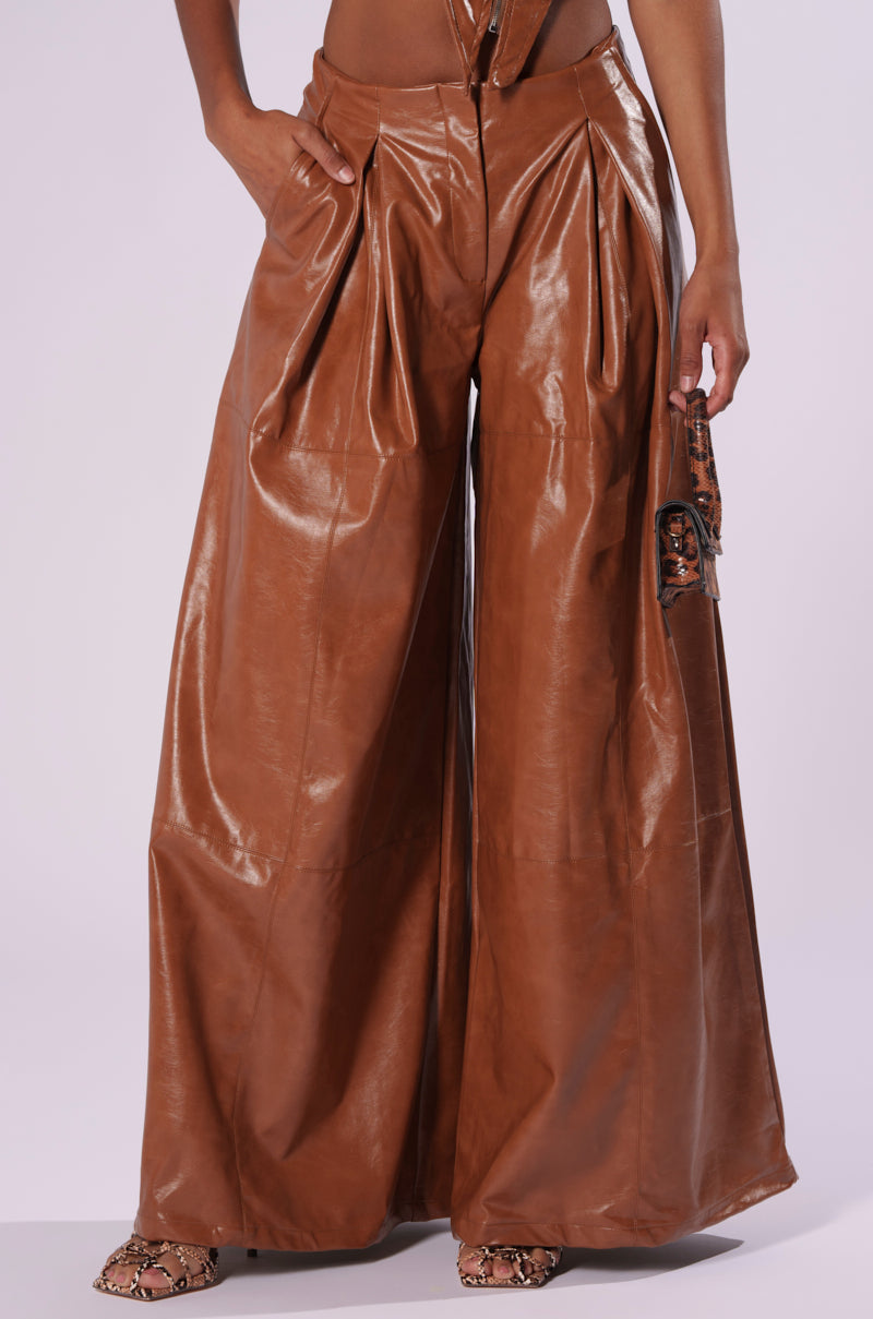A person is wearing the LOOK BACK AT IT WIDE LEG TROUSER, featuring high-waisted, wide-leg brown faux leather pants with pleats, complemented by a matching brown belt. They are also sporting snakeskin-patterned shoes. The image is cropped at the waist, emphasizing the stylish AKIRA trousers without showing the person's face.