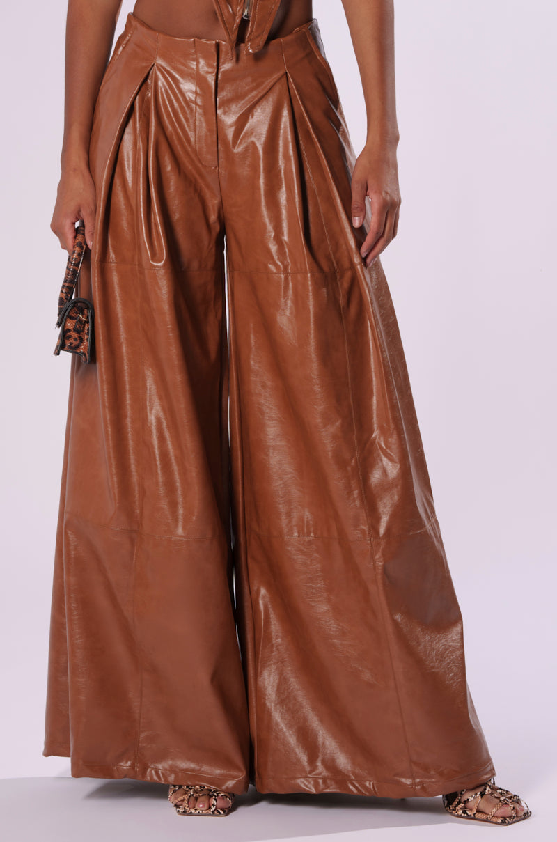 A person wearing high-waisted, wide-leg LOOK BACK AT IT WIDE LEG TROUSER in brown faux leather poses against a plain background. The trousers have a glossy finish and pleats near the waistband. The person also holds a small leopard-print bag and wears animal-print sandals.
