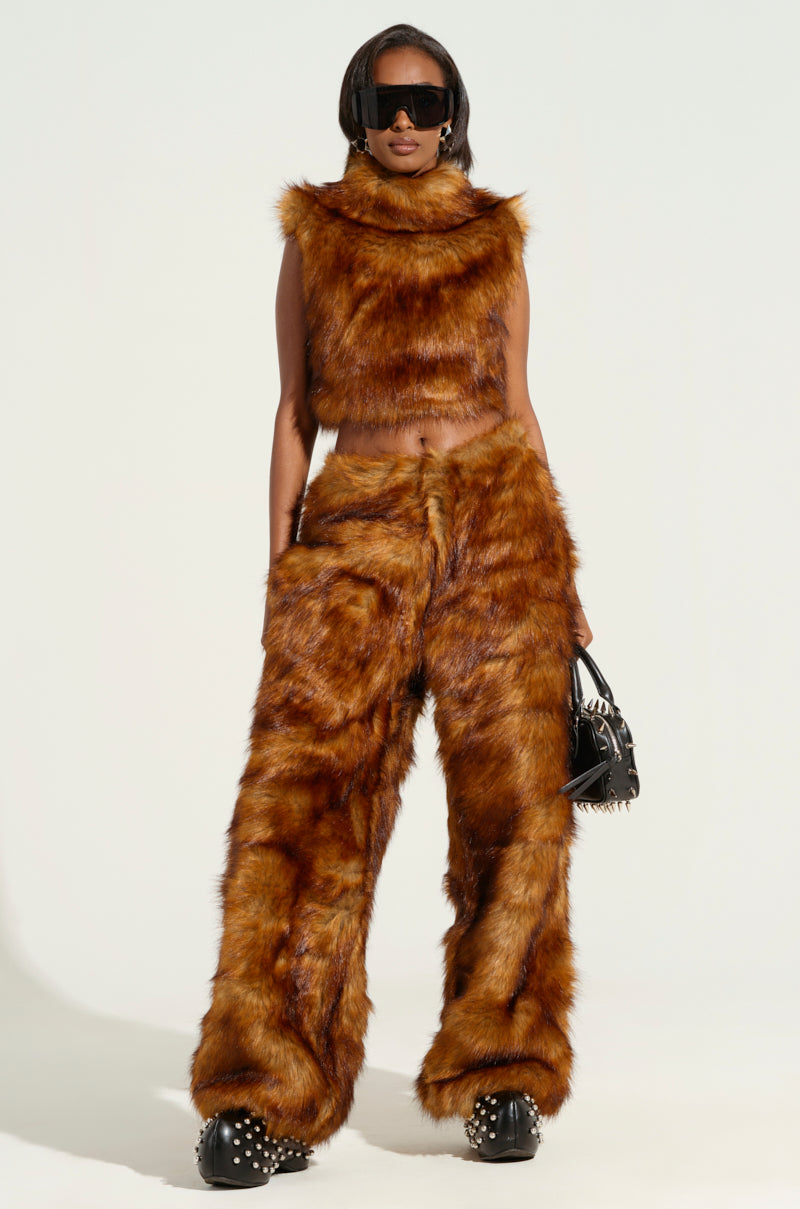 A person dressed in matching brown faux fur, featuring the JODIE WIDE LEG FASHION FUR PANT and a coordinating crop top, accessorized with large black sunglasses and a black studded bag. The ensemble is completed with black studded platform shoes. The background is plain light green.