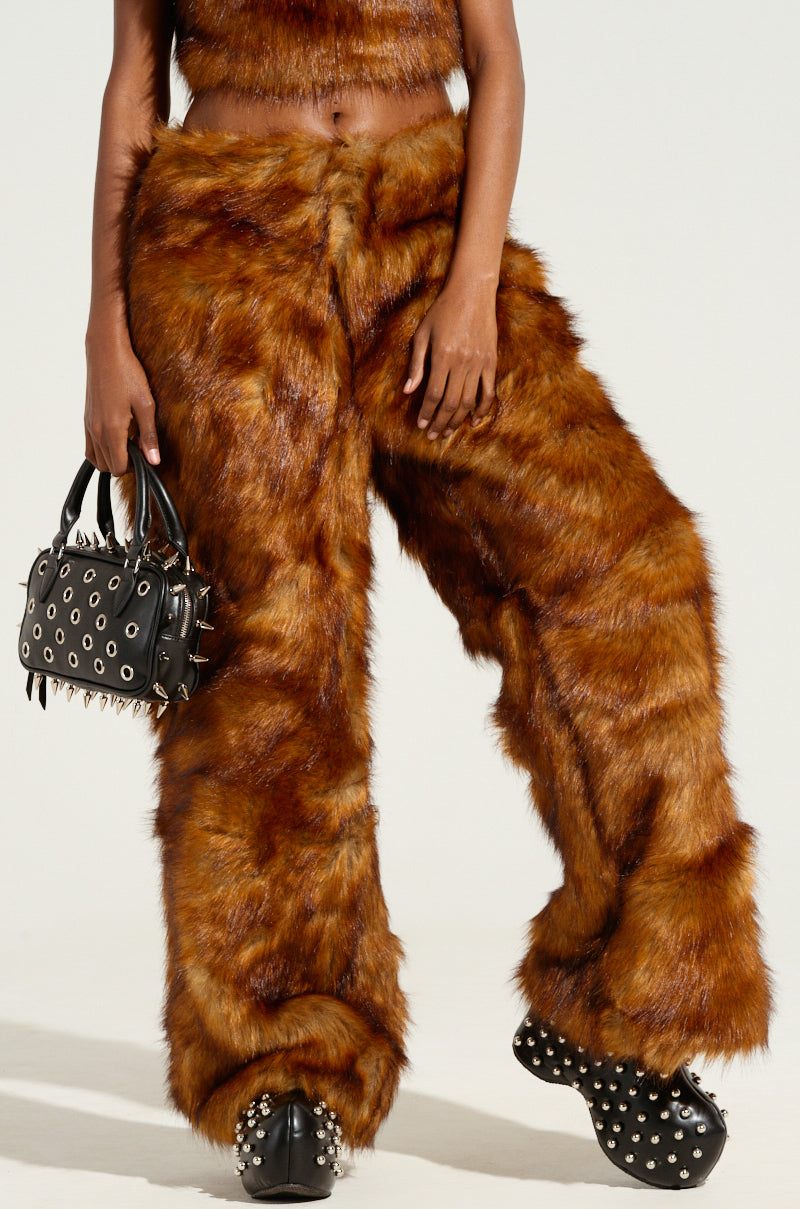 A person dressed in the JODIE WIDE LEG FASHION FUR PANT and a matching top carries a small black handbag embellished with spikes and studs. They complement the look with spiked black shoes against a plain, light-colored background.