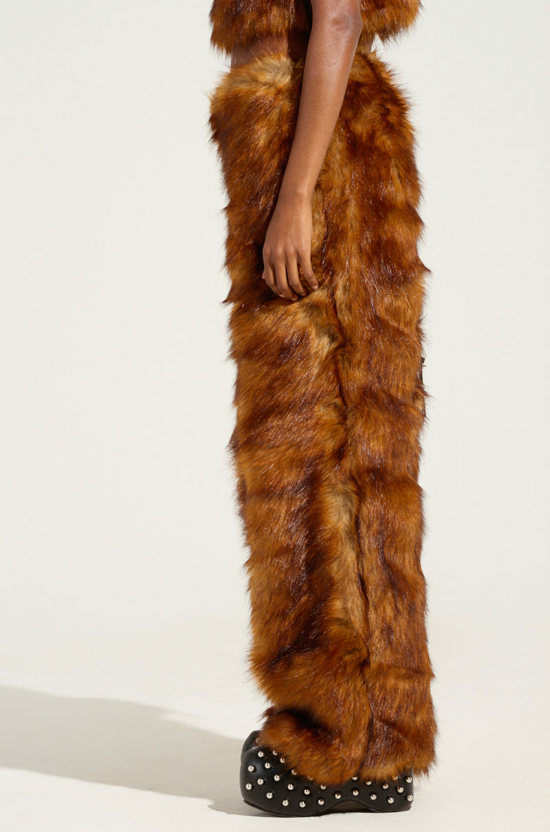 A person dressed in JODIE WIDE LEG FASHION FUR PANTS, distinguished by their brown faux fur material and wide leg design, stands against a plain background. These warm trousers fully cover their legs, complemented by black shoes adorned with white stud details. Their upper body is partially visible.