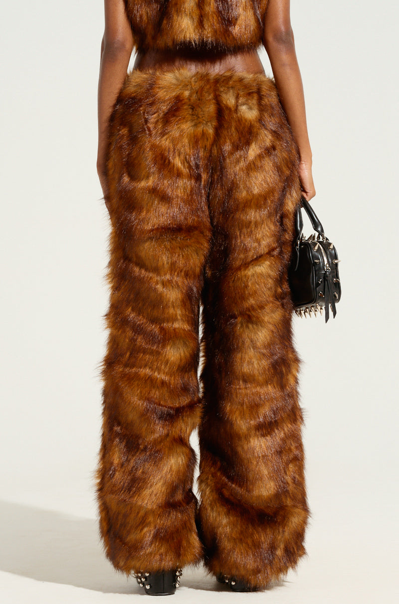 A person is standing, displaying the back view of their outfit made from brown faux fur, highlighting the JODIE WIDE LEG FASHION FUR PANT. They are holding a small black handbag adorned with metal studs against a plain, light-colored background.