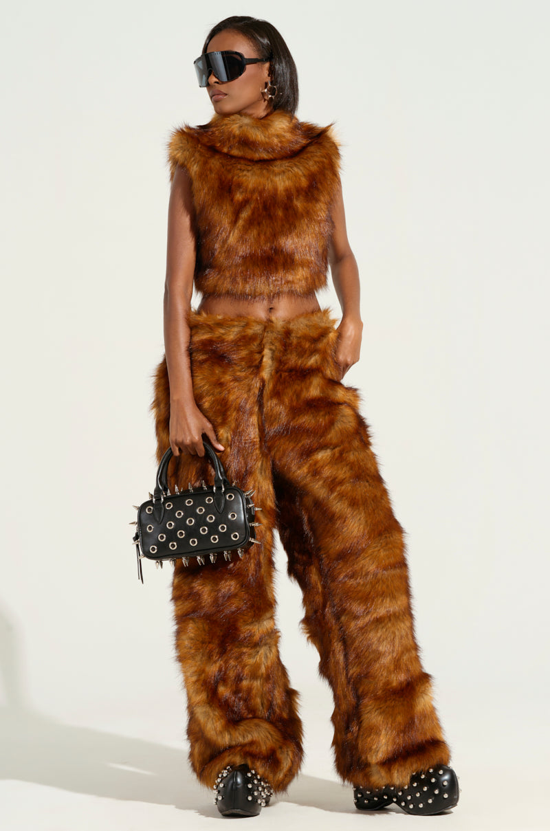 Dressed in a fashionable ensemble, an individual showcases the JODIE WIDE LEG FASHION FUR PANT in chic brown faux fur, with a matching sleeveless top. They accessorize with oversized black sunglasses and a studded black handbag, finishing their look with black shoes featuring metallic accents, set against a plain white background.