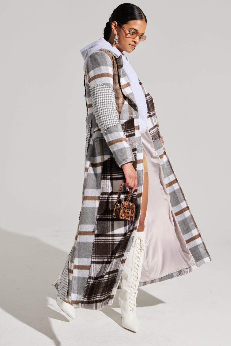 A person wearing the MIXED PLAID NOT FEELINGS PATCHWORK TRENCH over a light dress strides confidently. They accessorize with sunglasses, a small leopard-print purse, and white knee-high boots. The background is plain gray, enhancing the fashion-focused scene.