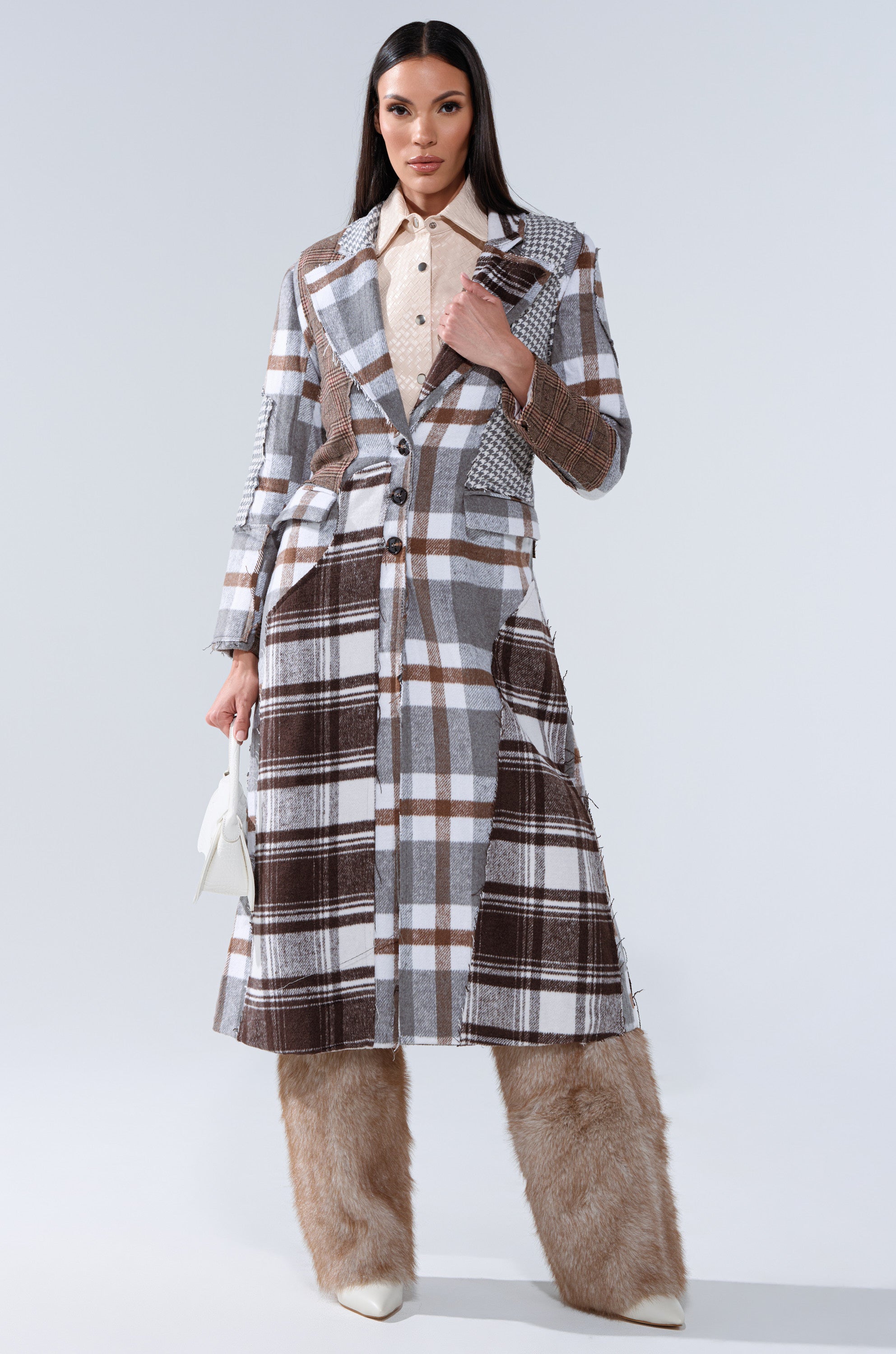 A woman stands confidently in the MIXED PLAID NOT FEELINGS PATCHWORK TRENCH over a beige shirt, tan faux-fur wide-leg pants, and white pointed shoes, holding a small white purse against a plain light gray background.