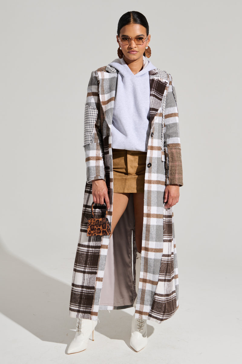 A person wearing sunglasses, the MIXED PLAID NOT FEELINGS PATCHWORK TRENCH with tortoiseshell button closures, a white hoodie, brown shorts, and white ankle boots stands against a plain background. They hold a small purse and have dark hair tied back.