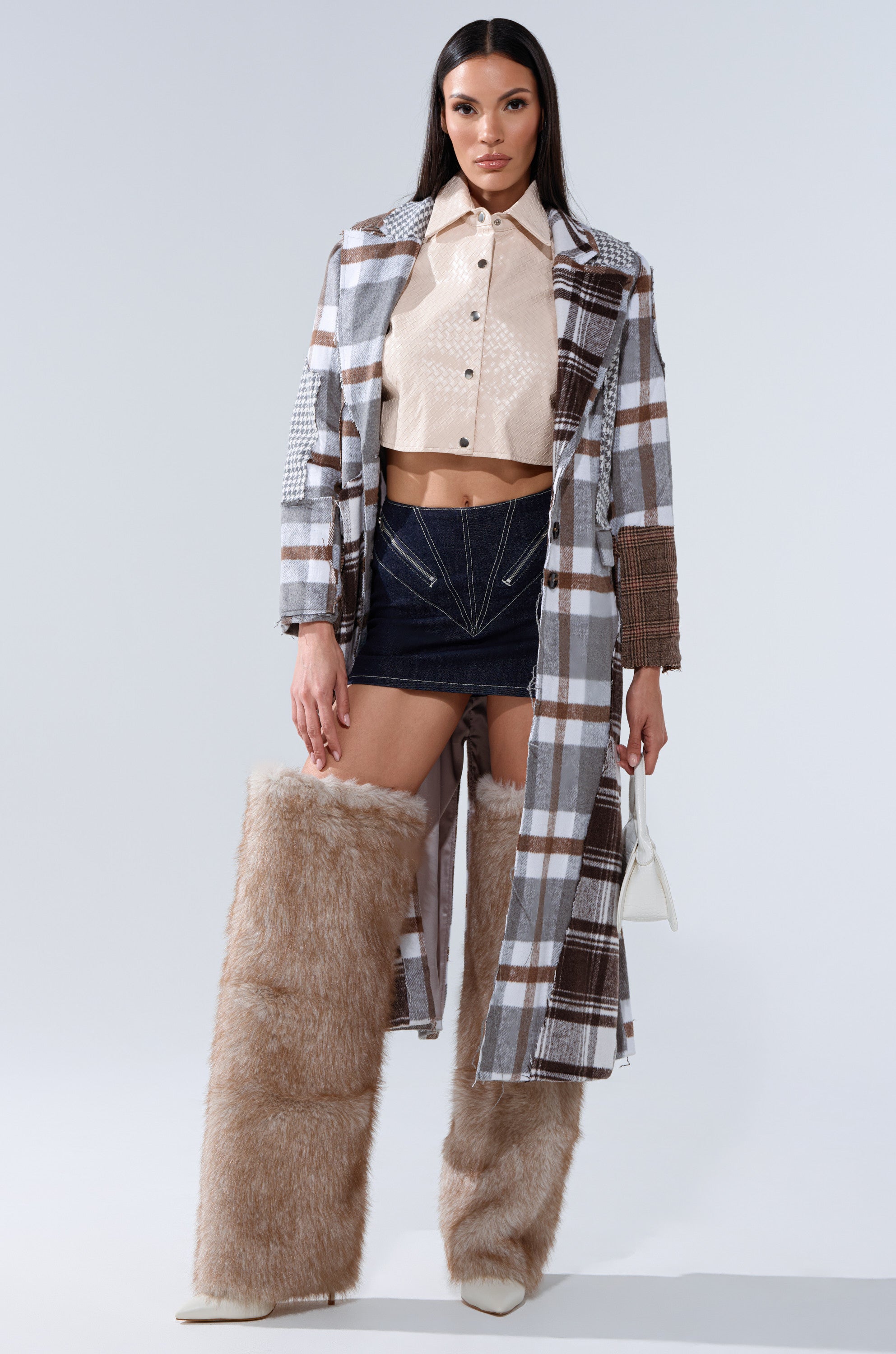 A woman wears the AKIRA Label MIXED PLAID NOT FEELINGS PATCHWORK TRENCH, a cream button-up crop top, short black skirt, tall light brown furry boots, and holds a small white handbag. She has straight dark hair.