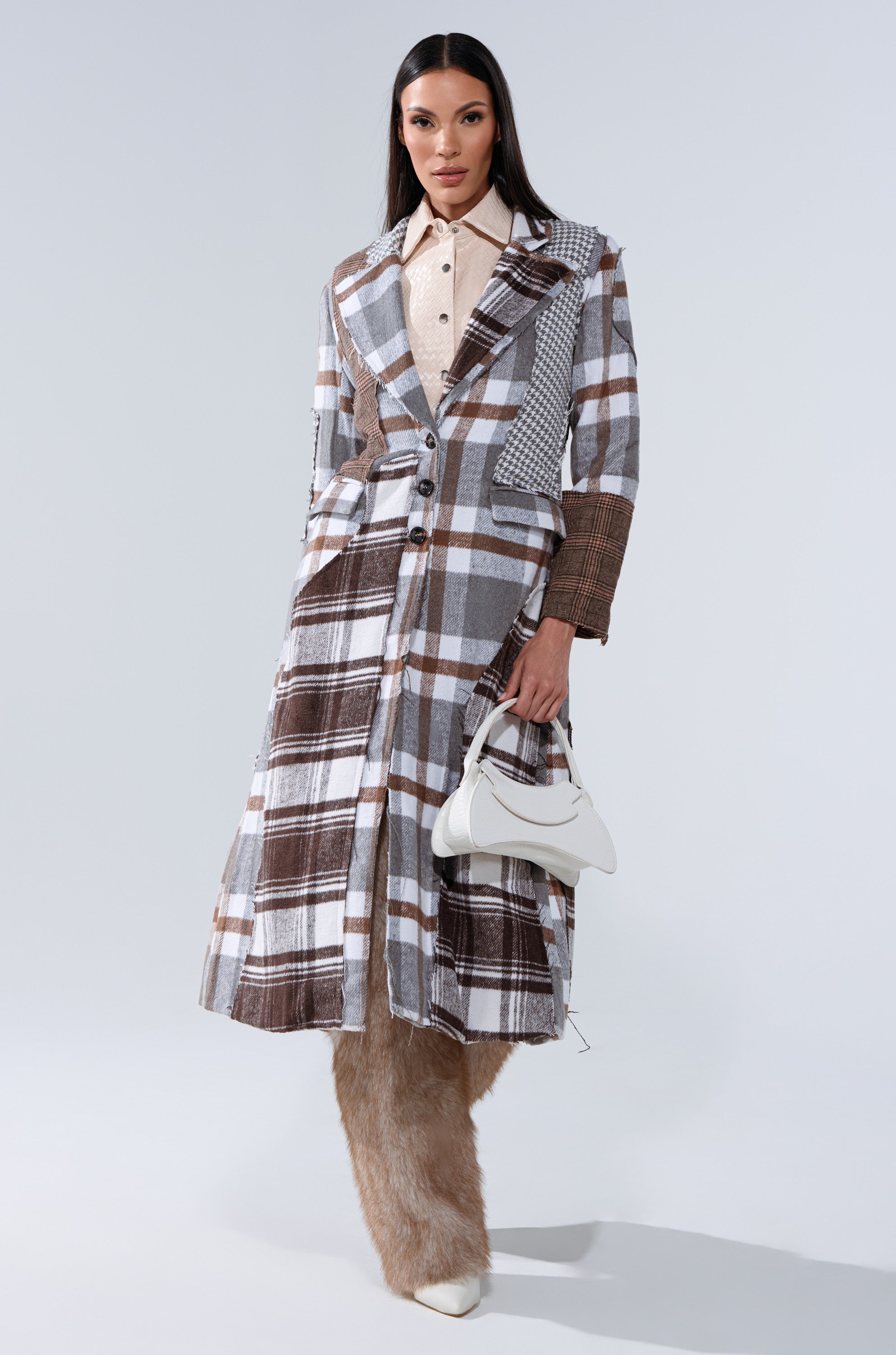 A woman poses in the MIXED PLAID NOT FEELINGS PATCHWORK TRENCH over a beige shirt, holding a small white handbag. She pairs wide furry tan pants with white pointed-toe shoes for a chic AKIRA look against a light background.